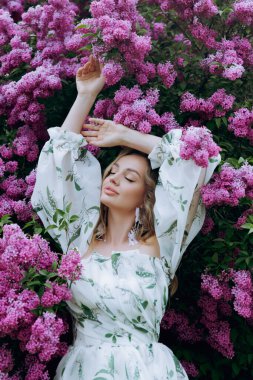 Beyaz elbiseli güzel, şık, tatlı sarışın. Leylak renkli bir fotoğraf çekimi için model olarak yeşil desenli. Onlar parkın yeşil bahçesinde onun lüks portrelerini yaptılar..