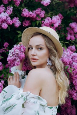 Beyaz elbiseli güzel, şık, tatlı sarışın. Leylak renkli bir fotoğraf çekimi için model olarak yeşil desenli. Onlar parkın yeşil bahçesinde onun lüks portrelerini yaptılar..