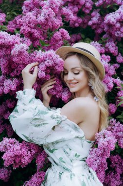 Beyaz elbiseli güzel, şık, tatlı sarışın. Leylak renkli bir fotoğraf çekimi için model olarak yeşil desenli. Onlar parkın yeşil bahçesinde onun lüks portrelerini yaptılar..