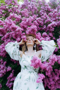 Beyaz elbiseli güzel, şık, tatlı sarışın. Leylak renkli bir fotoğraf çekimi için model olarak yeşil desenli. Onlar parkın yeşil bahçesinde onun lüks portrelerini yaptılar..