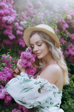 Beyaz elbiseli güzel, şık, tatlı sarışın. Leylak renkli bir fotoğraf çekimi için model olarak yeşil desenli. Onlar parkın yeşil bahçesinde onun lüks portrelerini yaptılar..