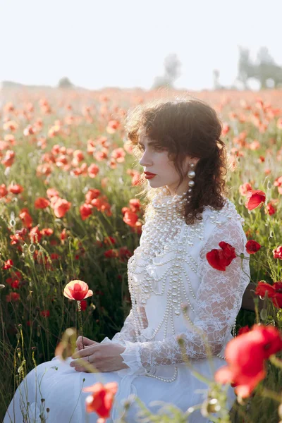 stock image A beautiful girl in a vintage white dress, as a model, at a fashion photo shoot in a poppy field at sunset.