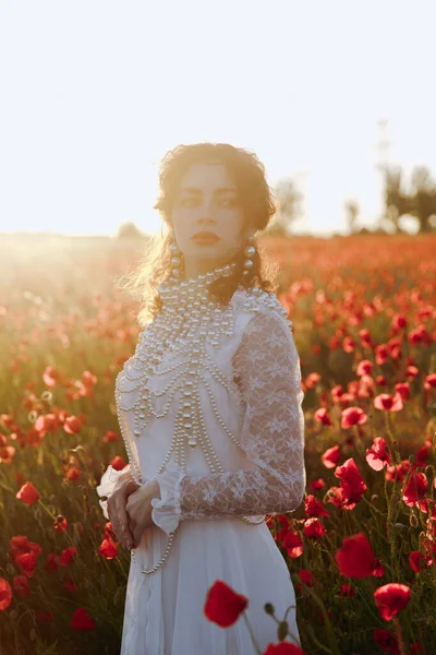 stock image A beautiful girl in a vintage white dress, as a model, at a fashion photo shoot in a poppy field at sunset.