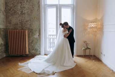 Damadın siyah takım elbiseli ve gelinin beyaz gelinlik içindeki ilk buluşması. Bir fotoğraf stüdyosunun ya da otelin içinde açık arkaplan balkonlu bir buket.