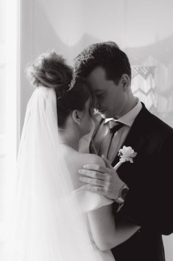 Damadın siyah takım elbiseli ve gelinin beyaz gelinlik içindeki ilk buluşması. Bir fotoğraf stüdyosunun ya da otelin içinde açık arkaplan balkonlu bir buket.