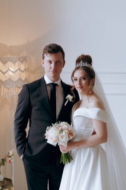Damadın siyah takım elbiseli ve gelinin beyaz gelinlik içindeki ilk buluşması. Bir fotoğraf stüdyosunun ya da otelin içinde açık arkaplan balkonlu bir buket.