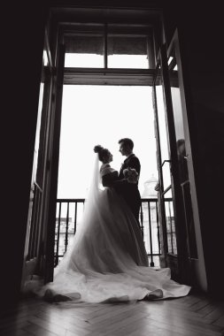Damadın siyah takım elbiseli ve gelinin beyaz gelinlik içindeki ilk buluşması. Bir fotoğraf stüdyosunun ya da otelin içinde açık arkaplan balkonlu bir buket.