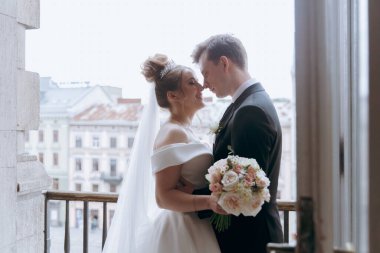 Damadın siyah takım elbiseli ve gelinin beyaz gelinlik içindeki ilk buluşması. Bir fotoğraf stüdyosunun ya da otelin içinde açık arkaplan balkonlu bir buket.