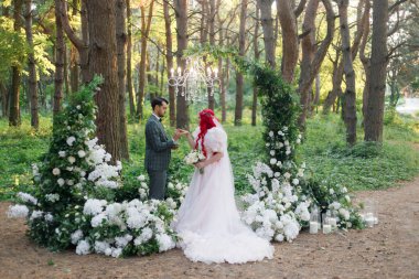 Güzel bir çift, yeşil takım elbiseli damat ve sıra dışı pembe gelinlikli gelin sonbaharda ormanda düzenlenen lüks bir açık hava seremonisinde poz veriyorlar.