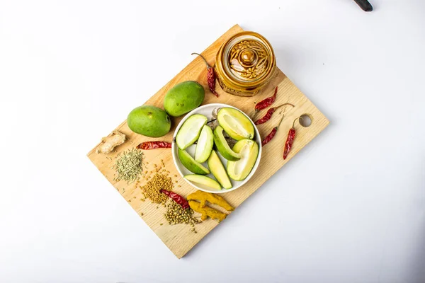 Indian mango pickle setup with green mango slices, jar and spices with white plate on wooden board