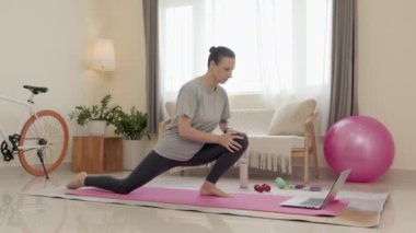 Woman in sportswear stretching on exercise mat at home and watching online fitness class on laptop