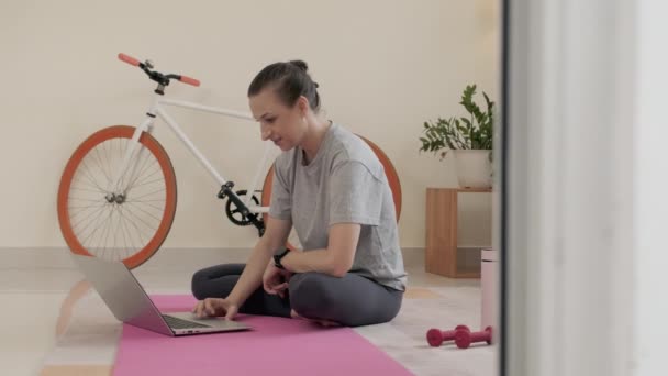 スポーツウェアの女性は自宅で運動マットに座って ラップトップ上でWebコールを介してオンラインフィットネスインストラクターと話す — ストック動画