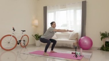 Full shot of woman doing squats and watching online video workout on smartphone held by tripod while exercising at home