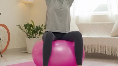 Tilt up shot of woman doing overhead tricep extension with dumbbells on exercise ball while training at home