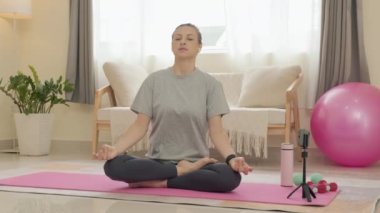 Woman sitting in lotus pose with eyes closed and hands in mudras while doing online yoga meditation at home with smartphone on tripod