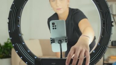 Female fitness instructor setting ring light with smartphone at home and checking time on wristwatch while preparing for filming video workout or giving online class