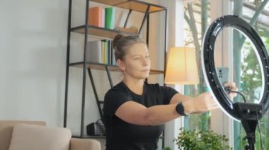 Female fitness influencer turning on ring light and smartphone camera while preparing for filming video workout or online streaming from home