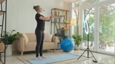 Full shot of female fitness instructor with headset showing exercises and speaking in mic in front of ring light with smartphone while filming sports vlog or training online from home