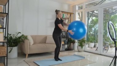 Full shot of female online trainer showing exercise with fitness ball in front of smartphone on tripod with circle lamp in living room at home