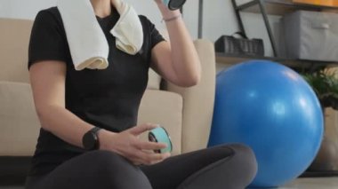 Tilt up shot of female fitness coach with headset drinking water from bottle and wiping face with towel while resting after giving online class at home