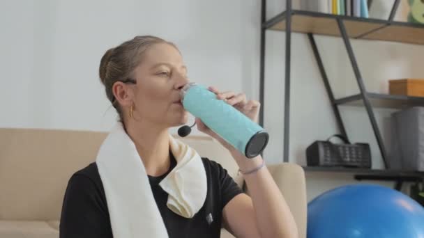 在家庭健身后放松时 用耳机喝水和毛巾擦拭脸的女性在线培训师的胸部照片 — 图库视频影像