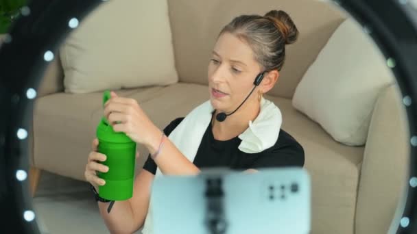 女性のフィットネスインフルエンサーの飲料水の選択的フォーカスショットとブログ上の製品を促進しながら スマートフォンカメラ上のスポーツシェーカーについて語る — ストック動画