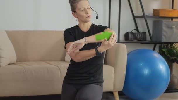 自宅でオンラインクラスを与えながら肘にテープを使用する方法を説明する女性コーチのショットを傾ける — ストック動画