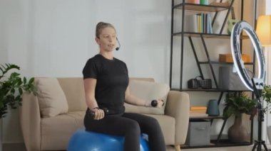Female coach showing dumbbell exercise on fitness ball in front of smartphone on tripod with circle lamp while giving live training session at home