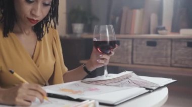 Young African American woman drawing fashion sketches and holding glass of red wine while working at home