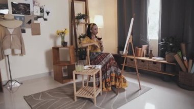 Relaxed African American woman sitting in living room at home, drinking red wine and enjoying her painting