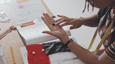 Close up shot of African American seamstress with measuring tape over her neck choosing buttons at desk in sewing workshop