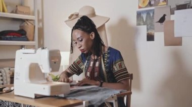 Young African American woman sitting at desk with sewing machine and working with fabric in tailor studio