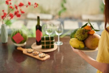 Cropped image of woman bringing plate with fresh fruits to dinner table