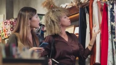 Medium shot of two young multiethnic women choosing garment and discussing it while going shopping together in clothes store