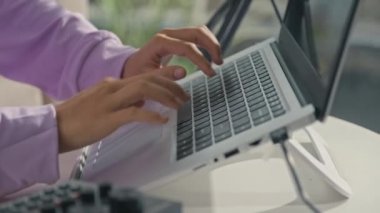 Cropped shot of hands of girl typing on laptop on stand while creating music at home studio with recording equipment