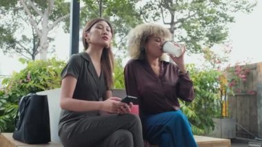Medium long shot of two young multiethnic women waiting for someone outside on street bench. One girl drinking coffee and checking time on wristwatch while her friend holding smartphone