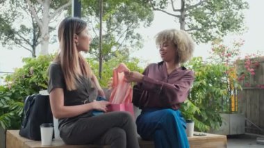 Medium long shot of two young female friends sitting on street bench with shopping bags and discussing new blouse