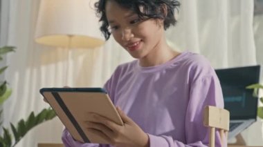 Tilt up shot of happy Asian gen Z girl using digital tablet and then putting on wireless earphones, watch music video and singing while resting at home