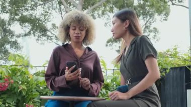 Medium shot of two young multiethnic women sitting in outdoor cafe on summer day and discussing social media post on smartphone