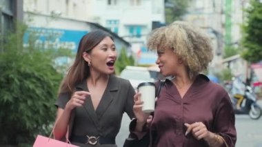 Medium slow motion shot of two young female friends smiling and discussing something while walking together in city with coffee cups and shopping bags