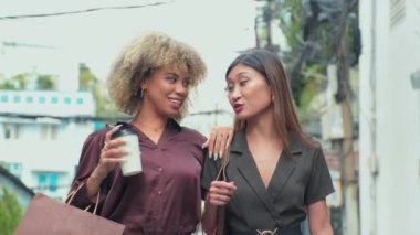 Waist up slow motion shot of two young female friends holding hands, chatting and laughing while walking together in city with coffee cups and shopping bags