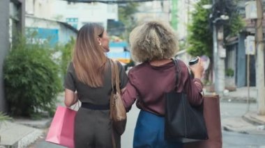Back view slow motion shot of two young women chatting while walking together on city street with coffee cups and shopping bags