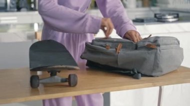 Tilt up shot of Asian gen Z girl zipping backpack and taking skateboard before leaving home in morning