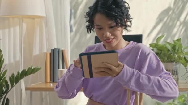 デジタルタブレットを使用して笑顔アジアのジェンZの女の子のショットを傾け その後 自宅で冷却しながら インターネット上で何かを見るために無線イヤフォンを置く — ストック動画