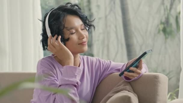 リラックスしたアジアのゲンZ女の子の中ショット自宅でソファに座って ワイヤレスヘッドフォンでスマートフォンで音楽を聴く — ストック動画