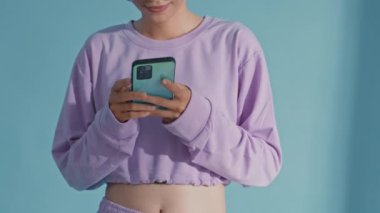 Tilt up shot of Asian gen Z girl in very peri outfit and trendy sunglasses typing on smartphone and posing for camera with smile against blue background in studio