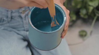 Close up impersonal shot of hands of girl stirring blue paint in can with paintbrush