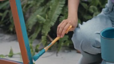 Cropped handheld camera shot of hands of girl painting old stool with blue color while restoring furniture outdoors on summer day