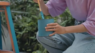 Close up tilt down shot of hands of girl painting stool with blue color while restoring old furniture outdoors on summer day