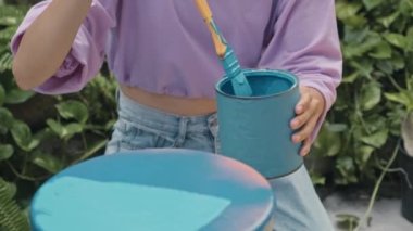 Cropped impersonal shot of girl painting old stool with blue paint while restoring furniture outdoors in backyard